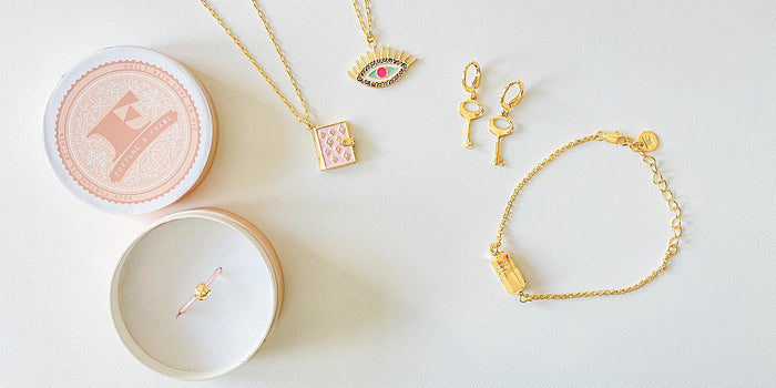 Fortune Cookie Ring (blush) in packaging, Stardust Book Locket (blush), Evil Eye Pendant (Wisdom), Mini Key of F Earrings, and Polygon Bracelet (gold) next to each other on a white background.